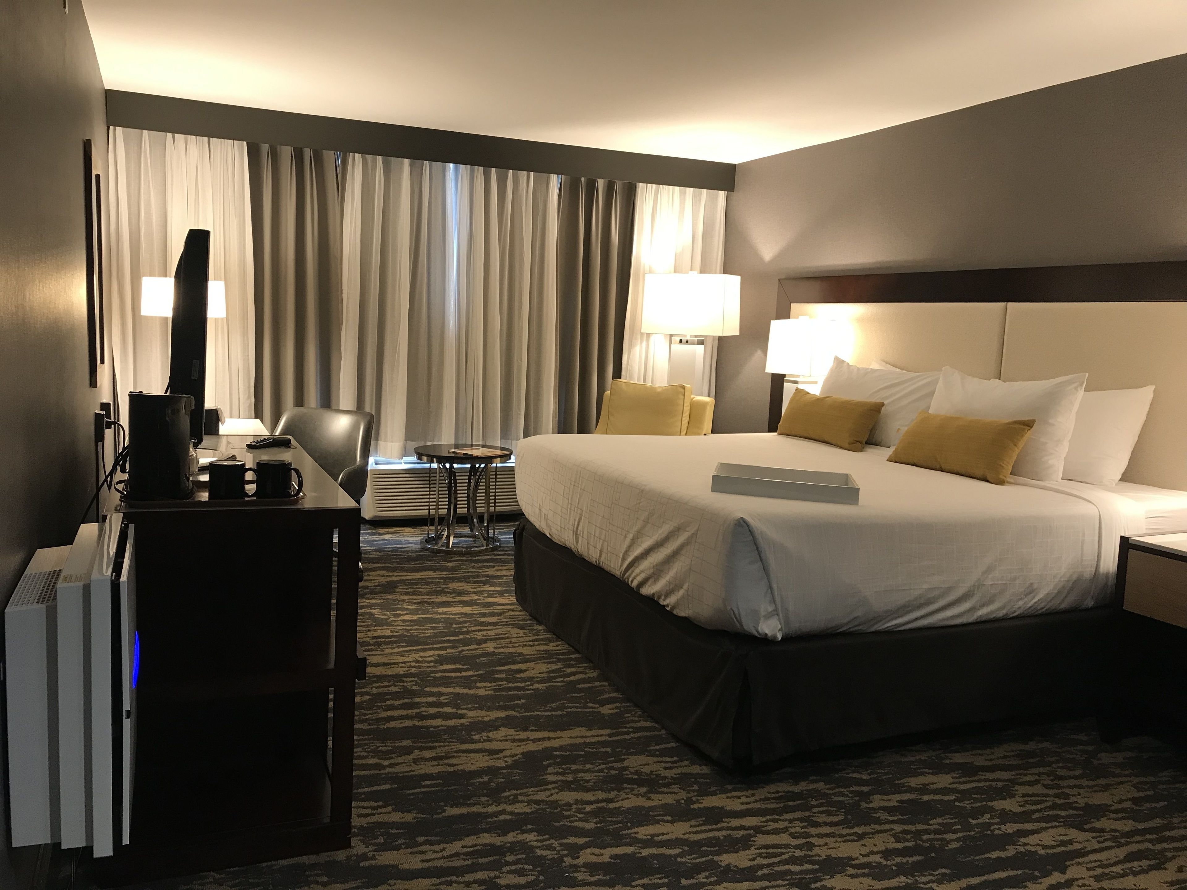 Una habitación de hotel con una cama grande, un escritorio con una computadora, una silla, una mesita y un ventanal con cortinas.