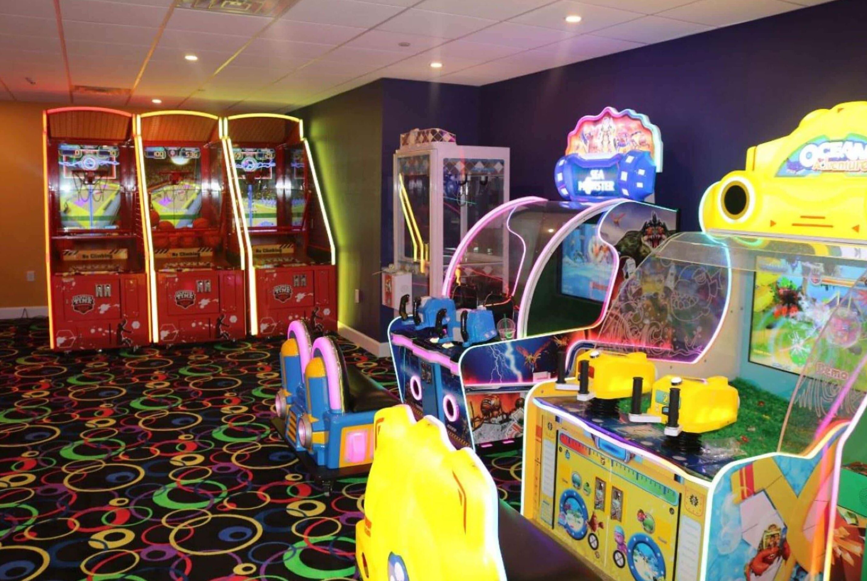 A brightly lit arcade room with various gaming machines, including basketball and racing games, and a colorful floor design.