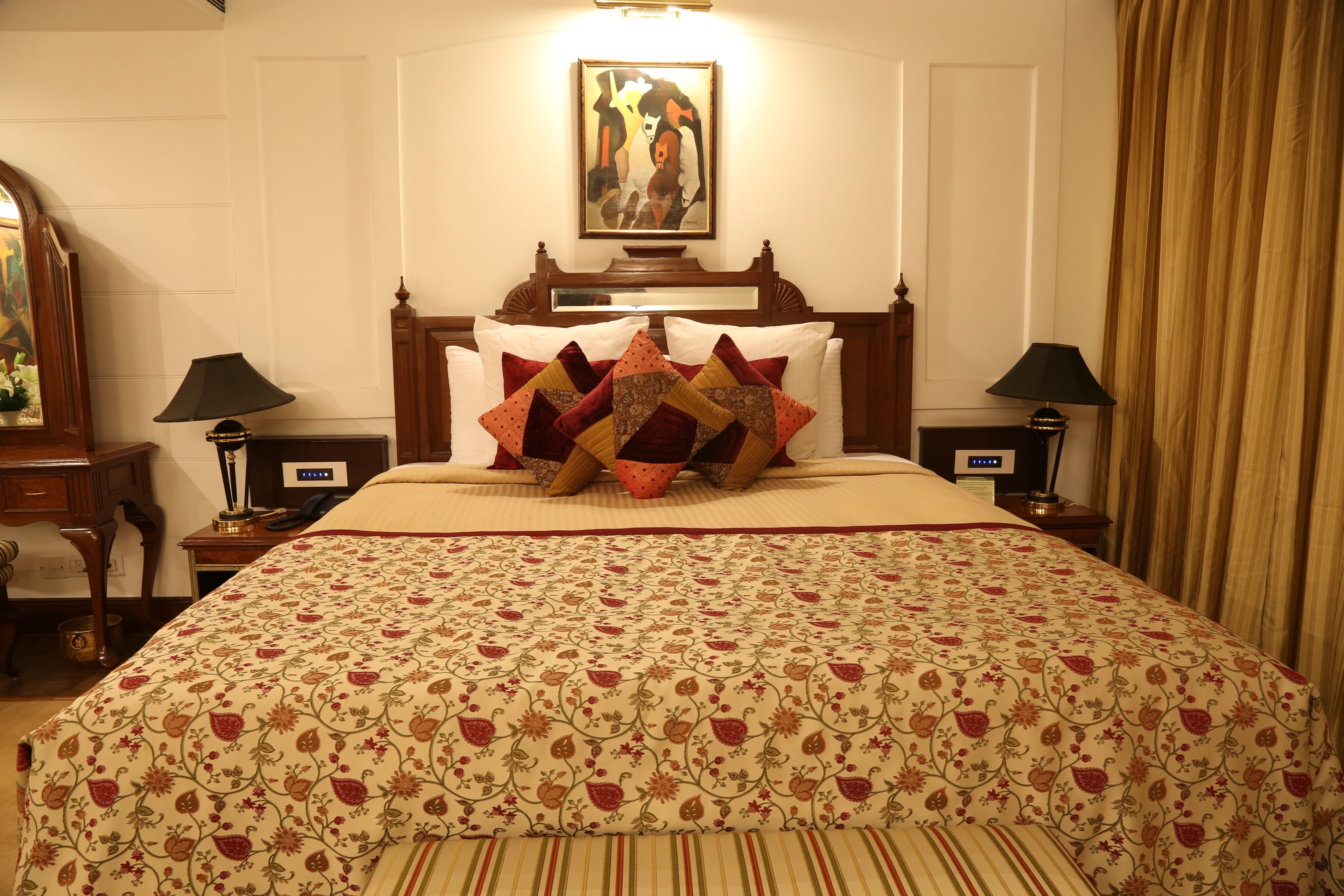 A neatly made bed with a floral-patterned bedspread and multiple decorative pillows. There is a wooden headboard, a framed artwork above the bed, and two bedside tables with lamps.