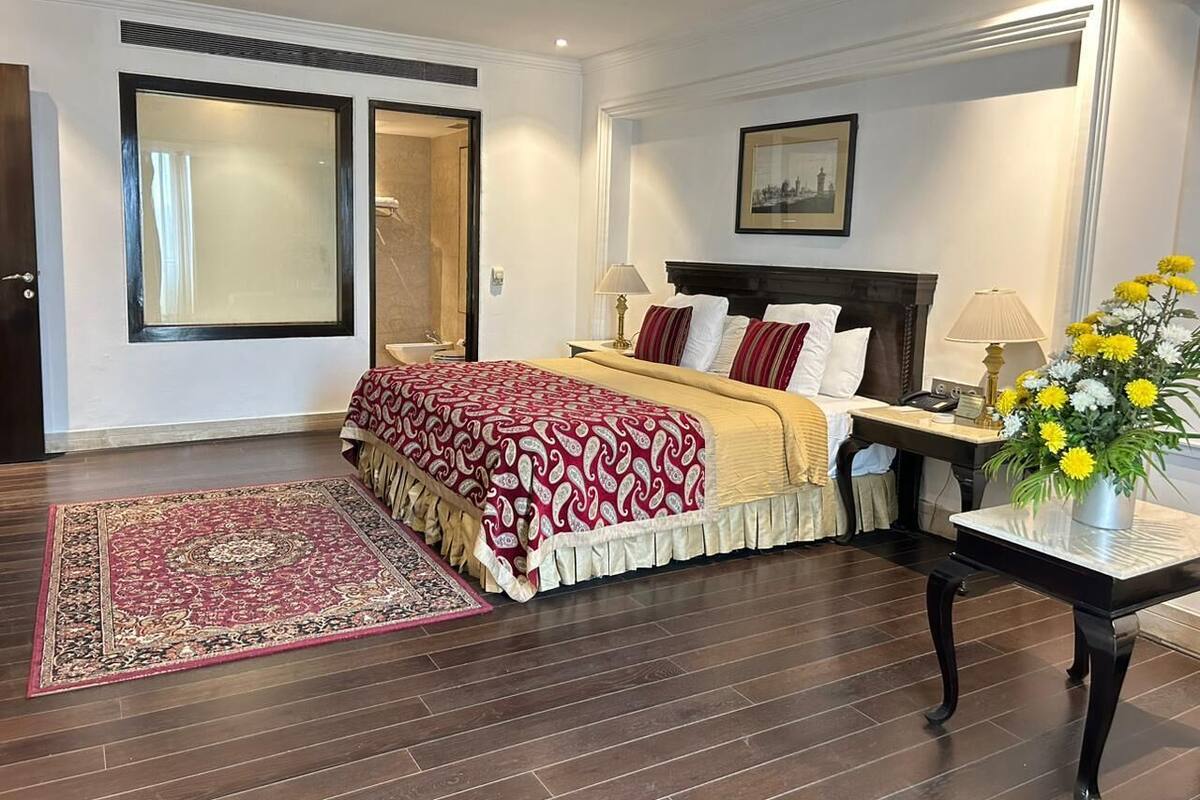 Une chambre avec un grand lit, une table de chevet avec une lampe, une commode, un tableau encadré au mur et une fenêtre avec des rideaux.