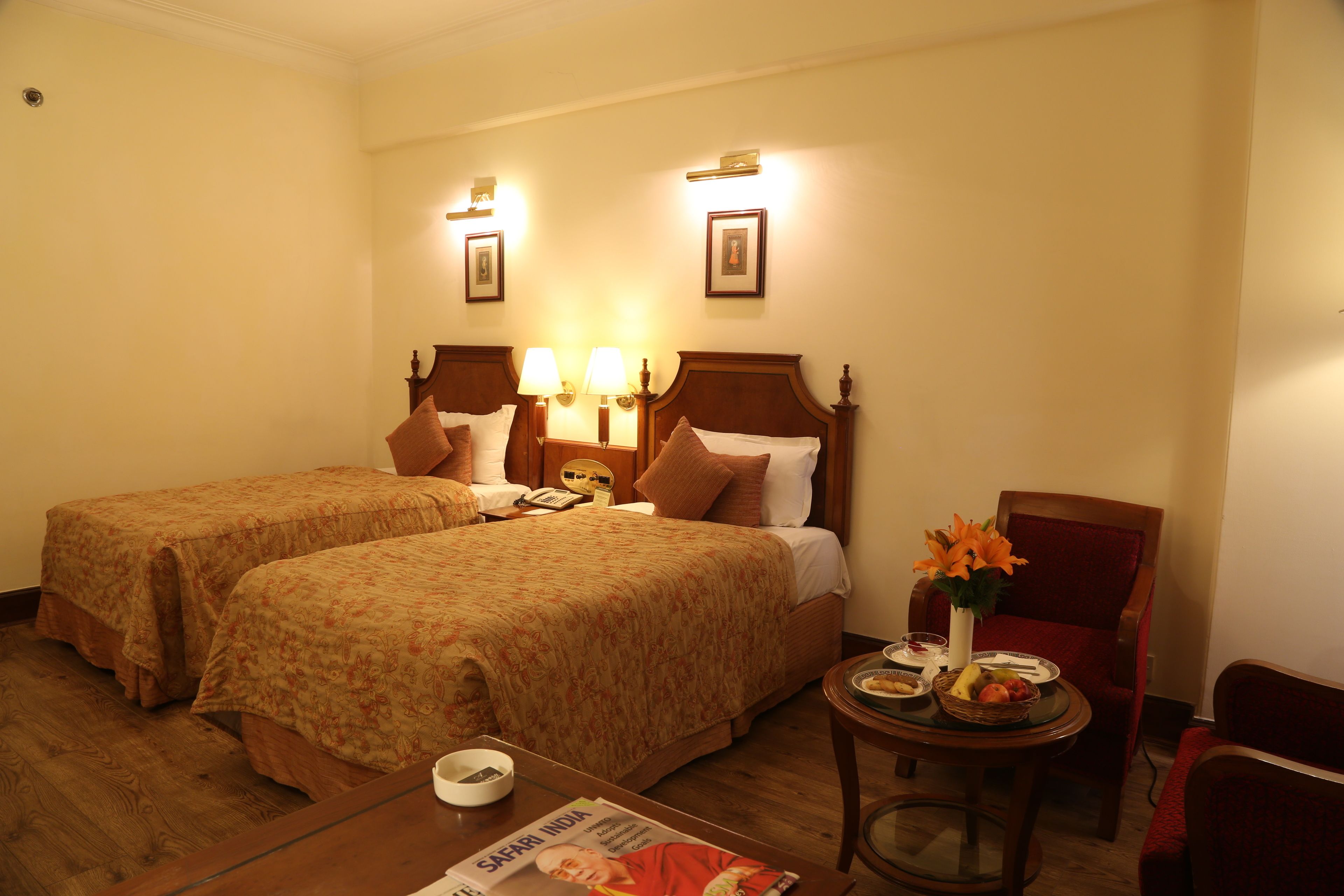 A hotel room with two beds, a small table with a magazine, a chair, and a small table with a fruit basket and a vase of flowers.