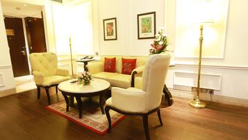 A well-lit living room with a yellow sofa, a white armchair, a round coffee table, and a floor lamp. There are framed pictures on the wall and a door leading to another room.