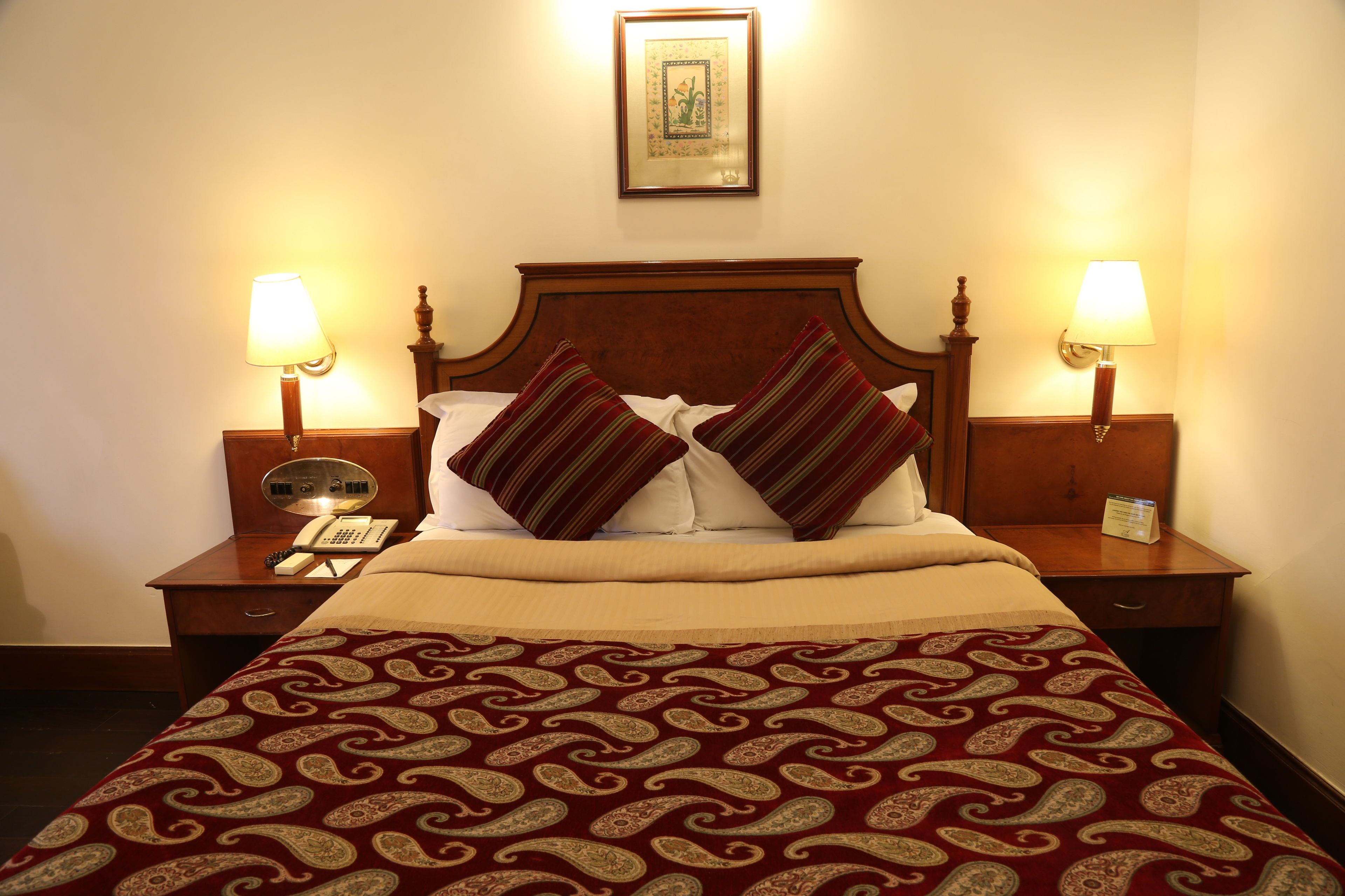 A neatly made bed with a patterned bedspread, two bedside lamps, a nightstand with a telephone, and a framed picture on the wall.