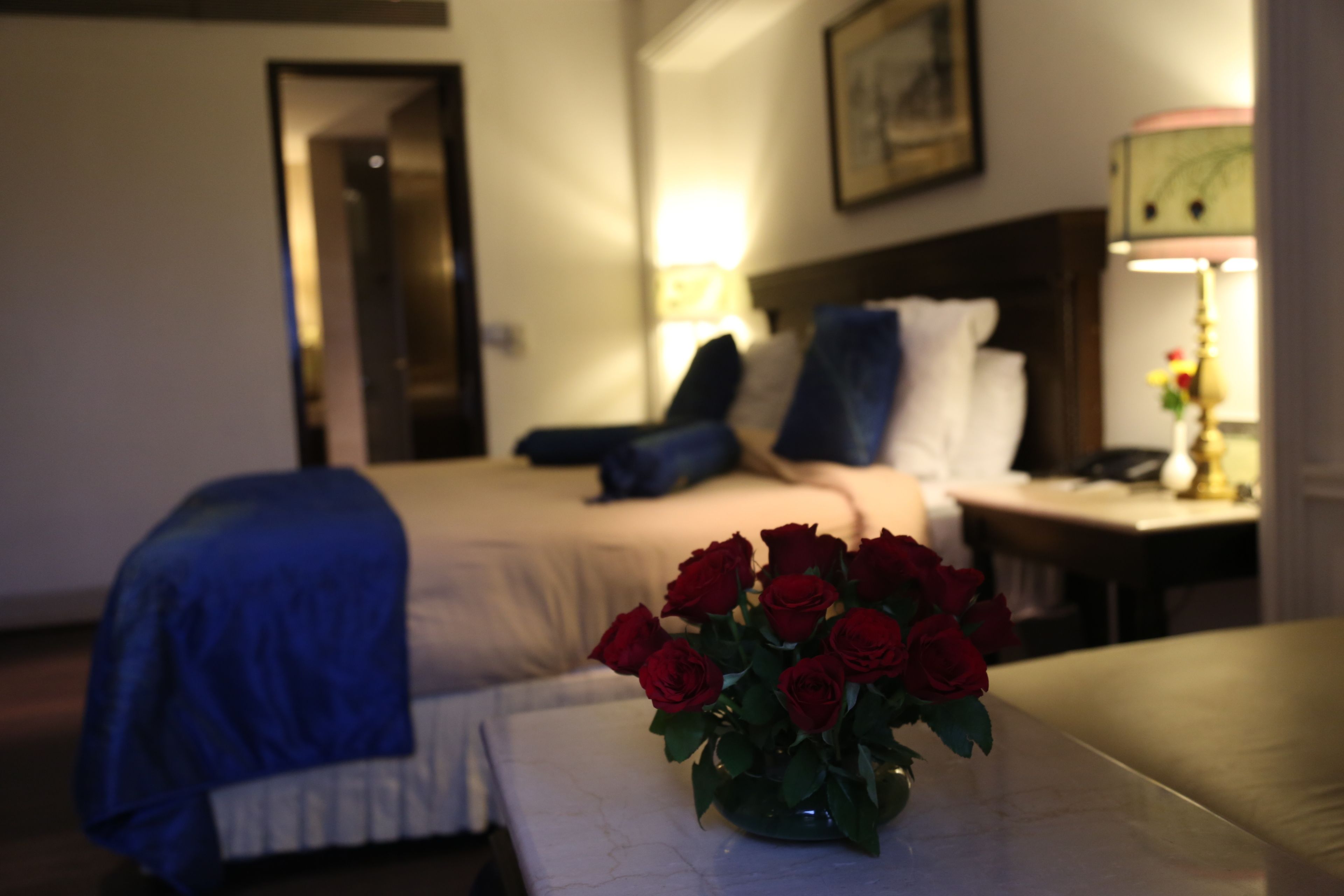 Una camera d'hotel con un letto, un comodino con una lampada e un vaso con rose rosse su un tavolo.