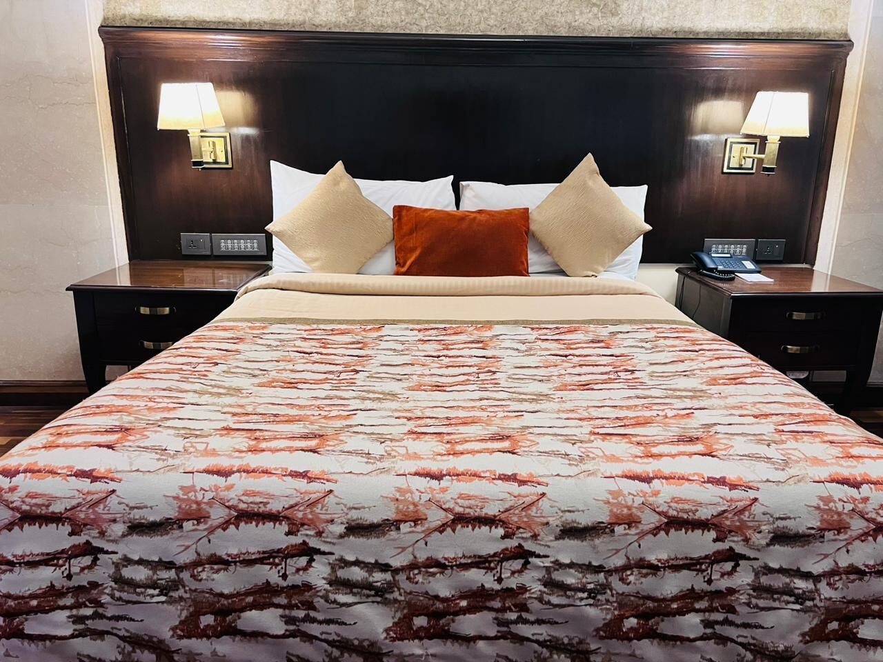 Una cama bien hecha con una colcha estampada, dos lámparas de noche y una mesita de noche con un teléfono y una pequeña mesa.