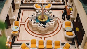 Un hall d’hôtel doté d’une fontaine centrale, de plusieurs espaces de détente avec des coussins jaunes et d’un comptoir d’enregistrement.