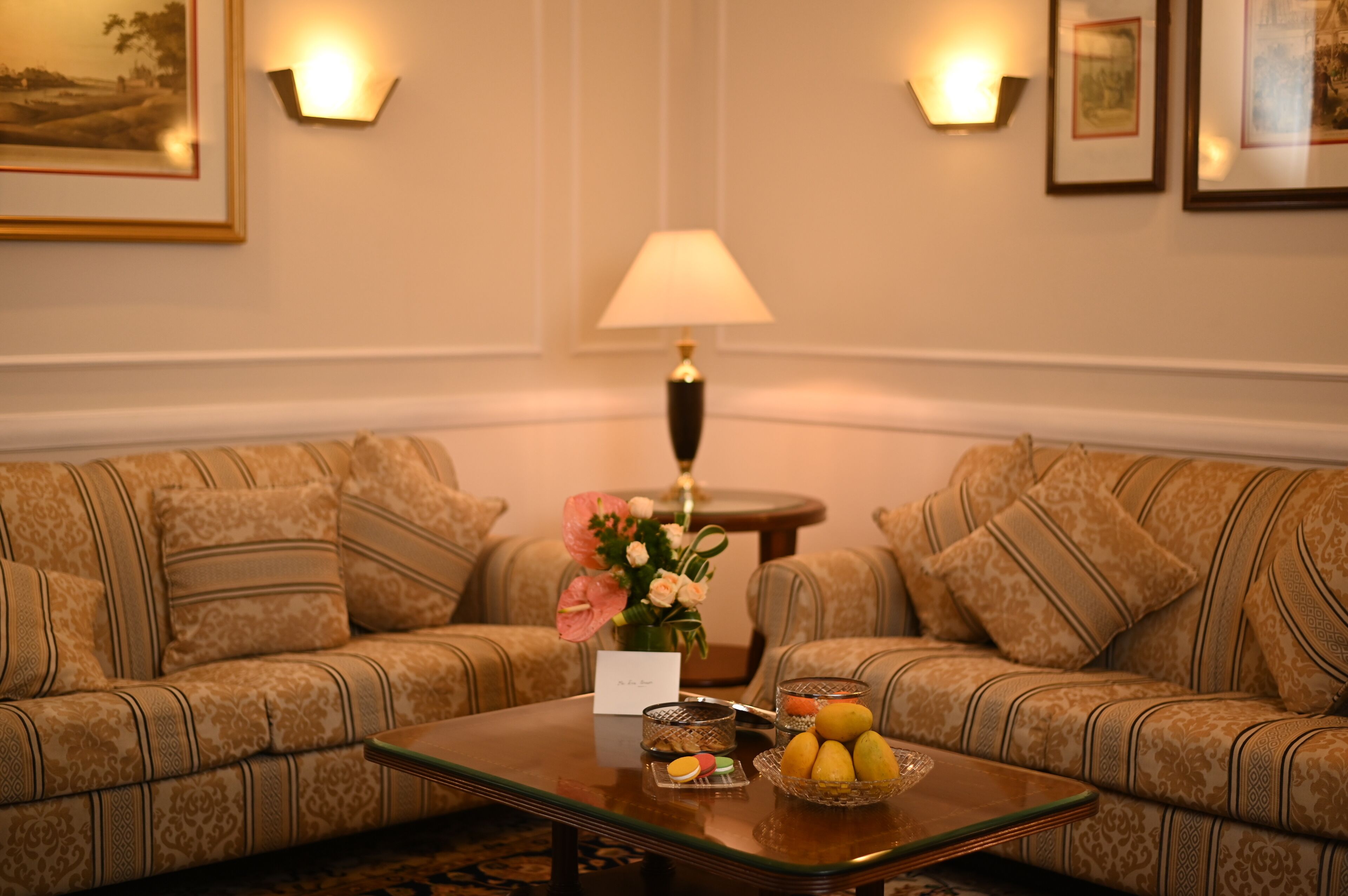 A room with two patterned sofas facing each other, a small round table with a lamp, a vase with flowers, and a fruit bowl.