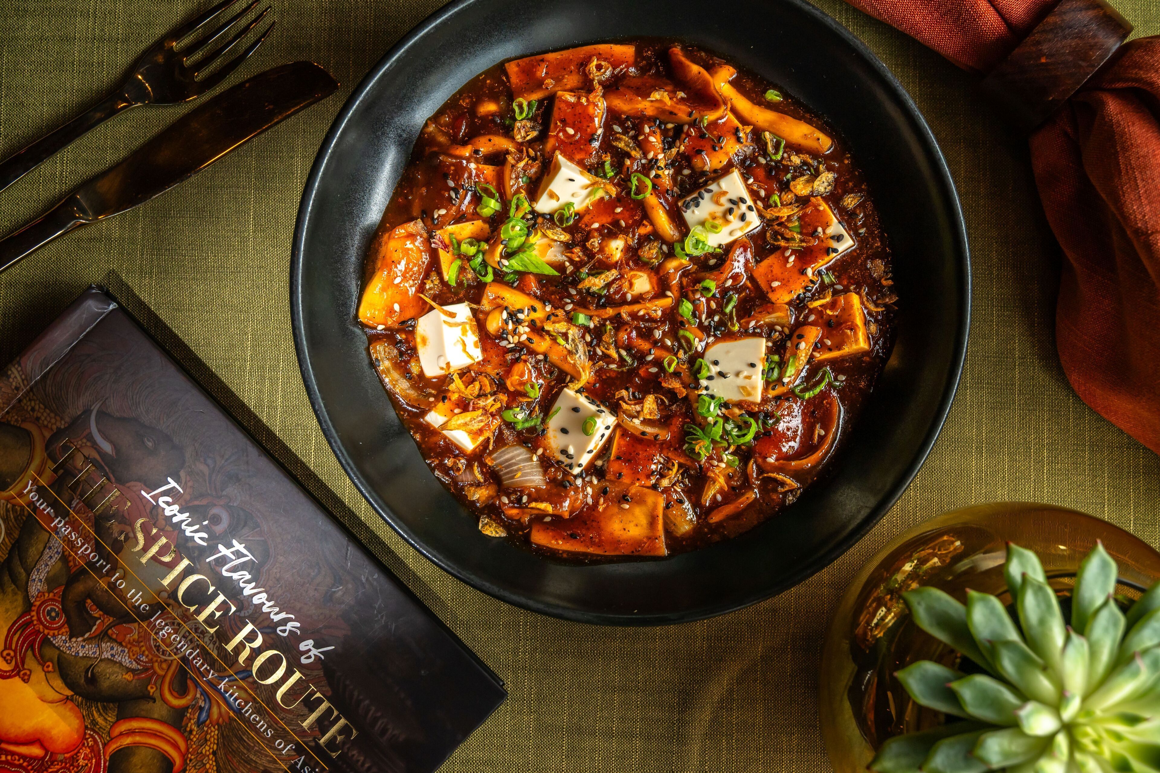 A plate of spicy tofu dish with a book titled 'Tonic Flavours of Spice Route' beside it.