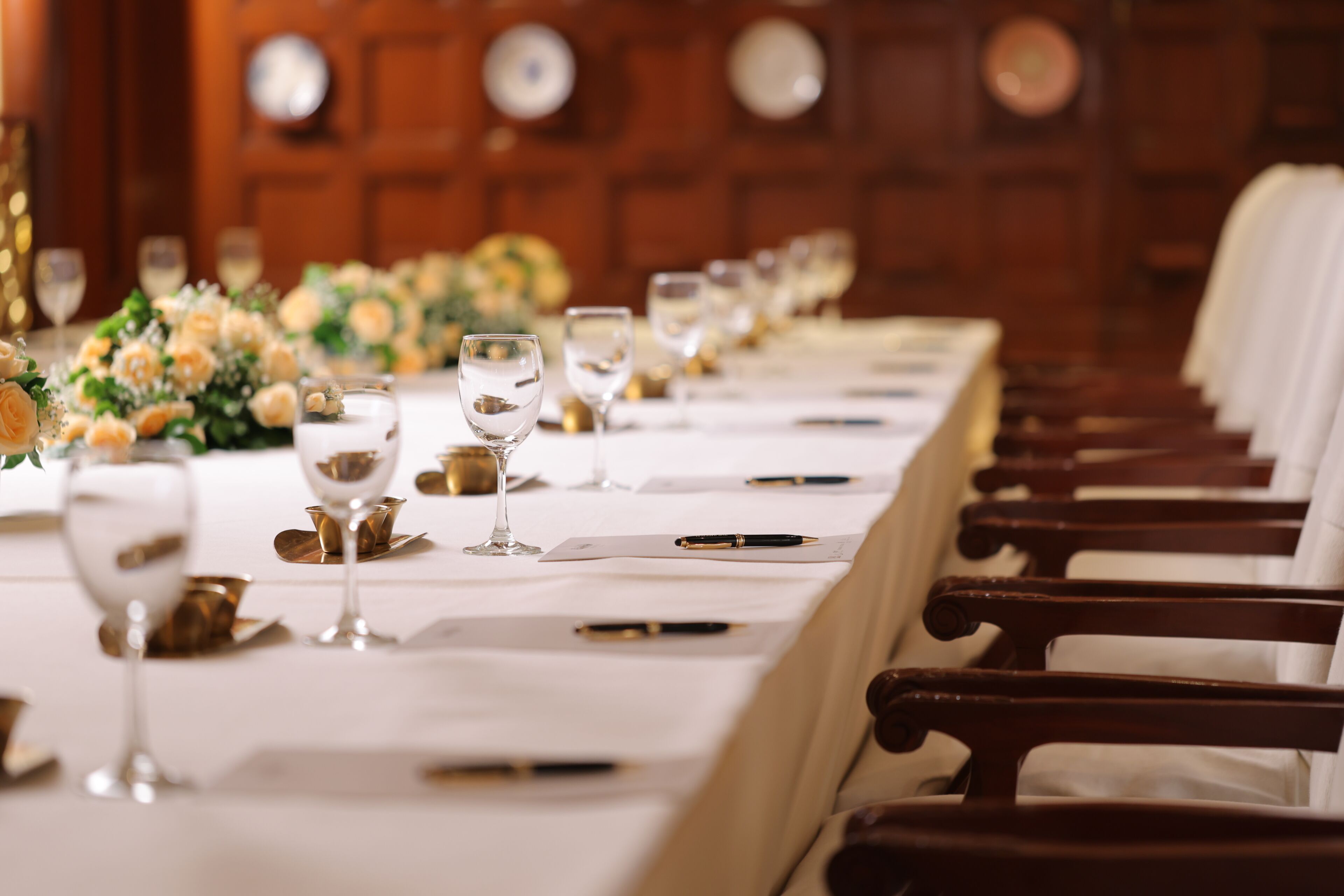 Una larga mesa de comedor con sillas, vasos y un centro de mesa floral.
