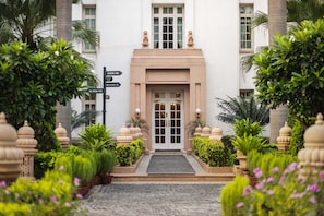 Entrada de um edifício com portas duplas, um pequeno jardim com vasos de plantas e um poste de sinalização.