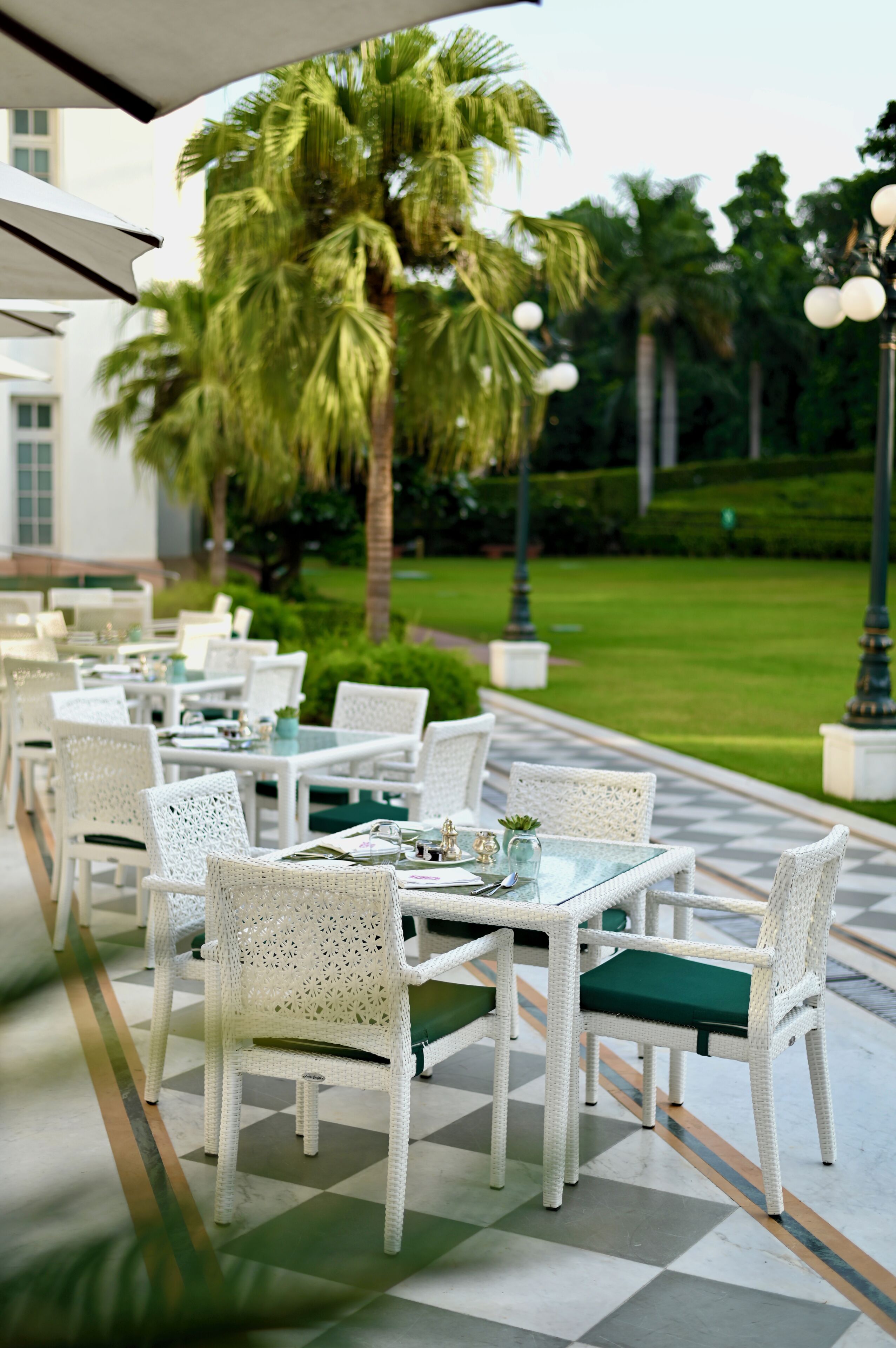 An outdoor dining area with white wicker chairs and tables, palm trees, and a well-maintained lawn.