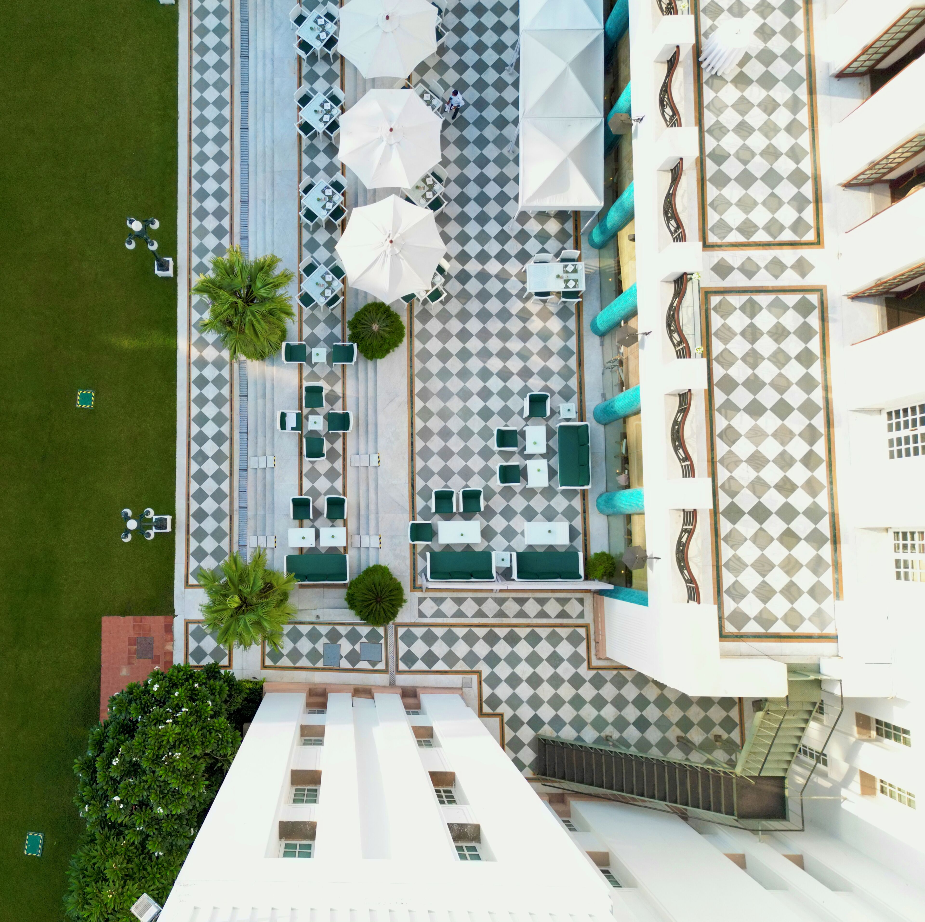Vue aérienne d’un hôtel dont le sol présente un motif à carreaux, avec des espaces de repos en plein air dotés de parasols et un escalier menant à un étage supérieur.