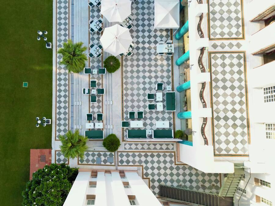 Vue aérienne d’un hôtel dont le sol présente un motif à carreaux, avec des espaces de repos en plein air dotés de parasols et un escalier menant à un étage supérieur.