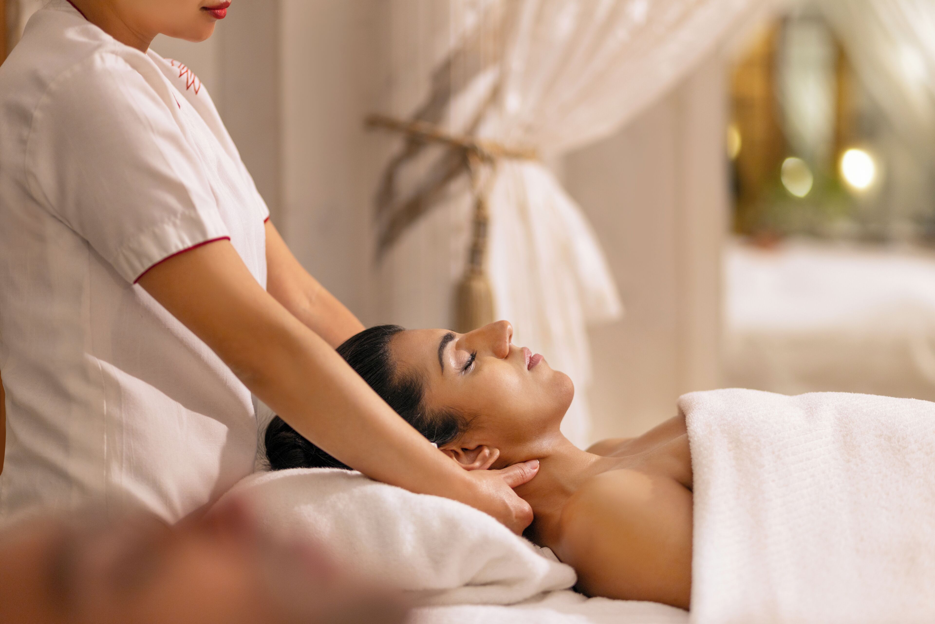 A person receiving a massage in a room with a bed, a white wall, and a curtain.