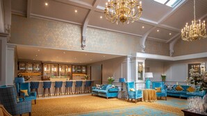 A spacious lobby with a bar area, blue upholstered seating, chandeliers, and patterned carpet. - Old Ground Hotel (Ennis)