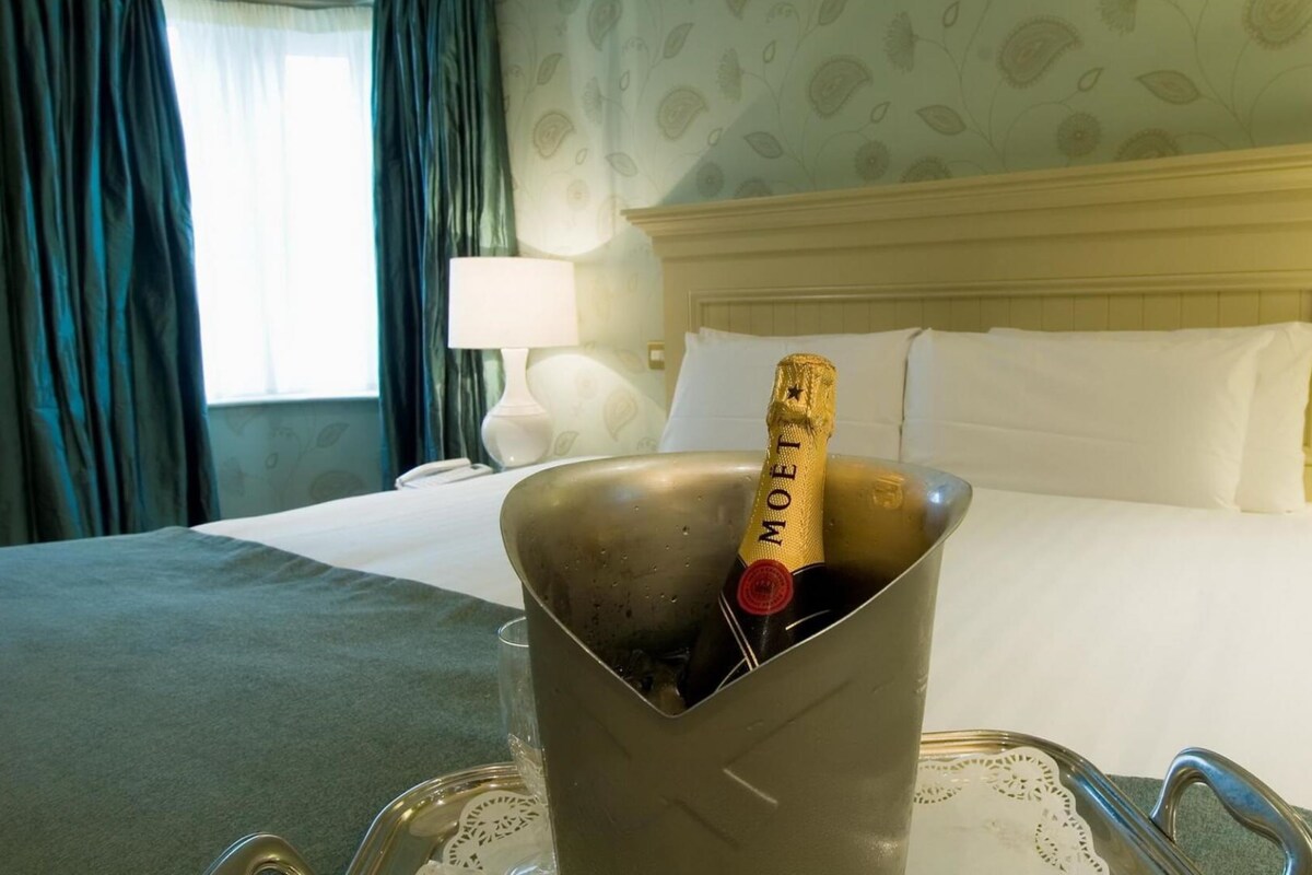 Et hotelværelse med seng, sengetøj og en champagneflaske i et glas på et fad.