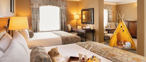 A hotel room with a large bed, a smaller bed, a desk, a chair, a mirror, a window with curtains, a yellow teepee, and a tray with cookies and a toy car.