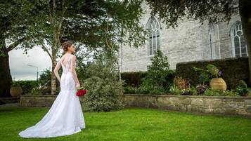 A bride in a white dress with lace details, standing in front of a stone building with arched windows.