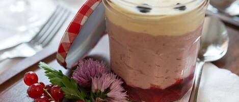 A layered dessert in a glass mug with a handle, garnished with red berries and a sprig of purple flowers, placed on a wooden table with a spoon and a fork beside it.
