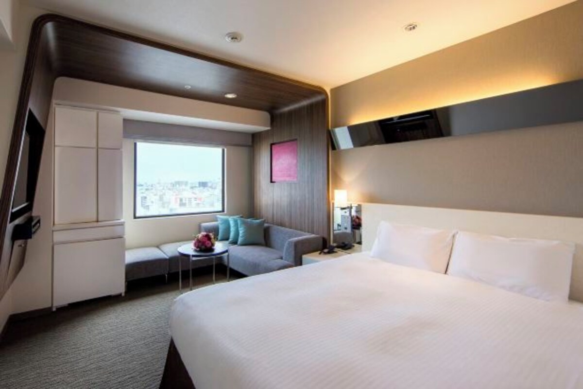 A modern hotel room with a large bed, a seating area with a small table, and a view of the city through a large window.