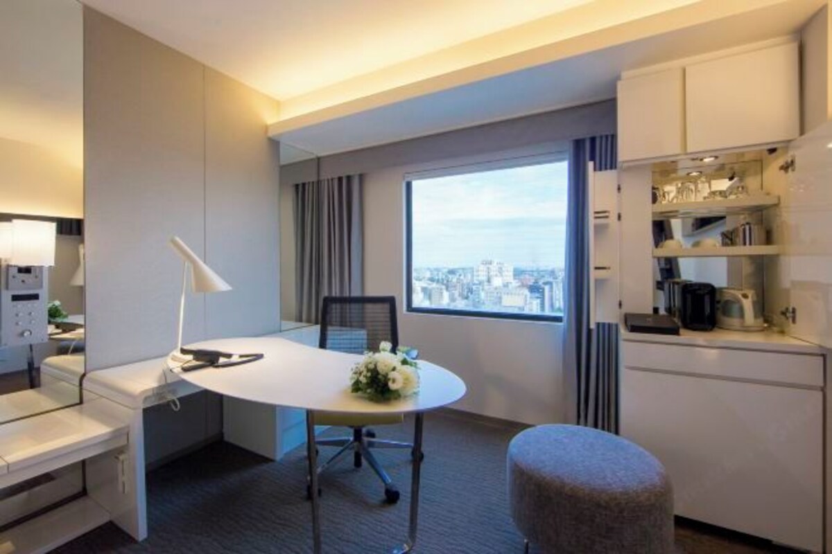 A hotel room with a large window offering a city view, a white desk with a lamp, a small kitchenette, and a comfortable seating area.