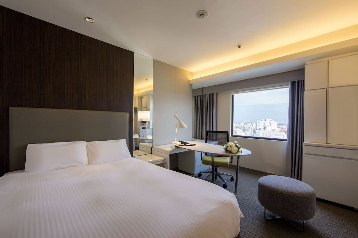 A hotel room with a large bed, a desk, a chair, and a view of the city.