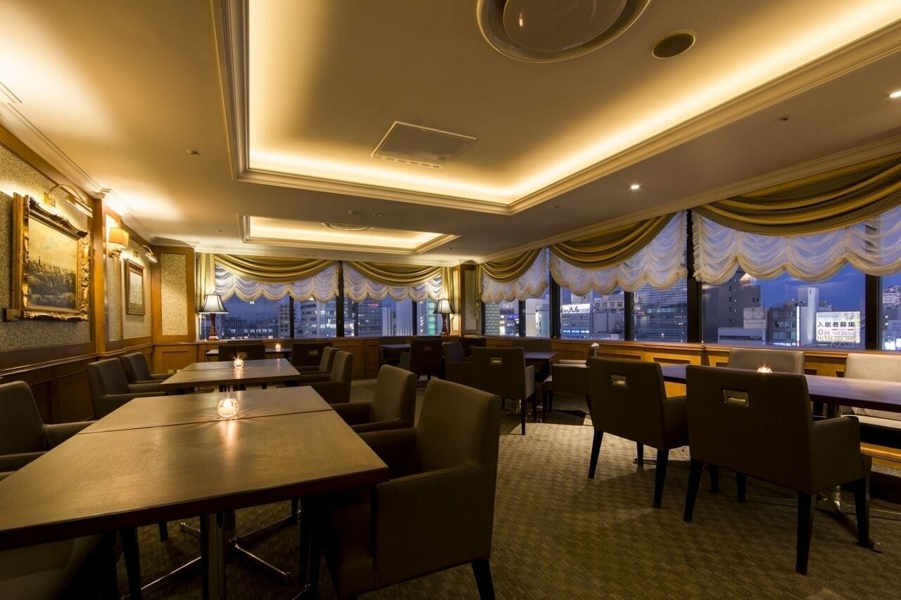 A hotel dining area with tables and chairs, large windows with curtains, and a city view.