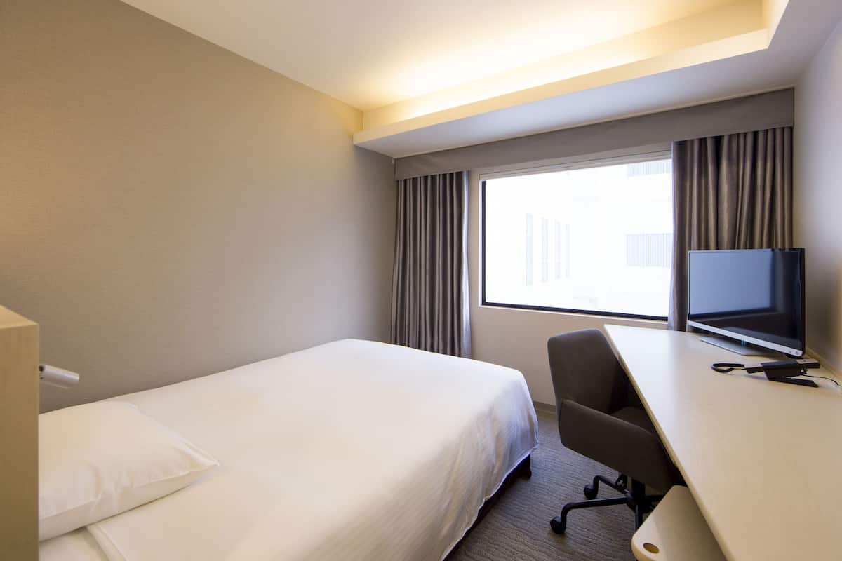 A hotel room with a single bed, a desk with a chair, a television, and a window with curtains.