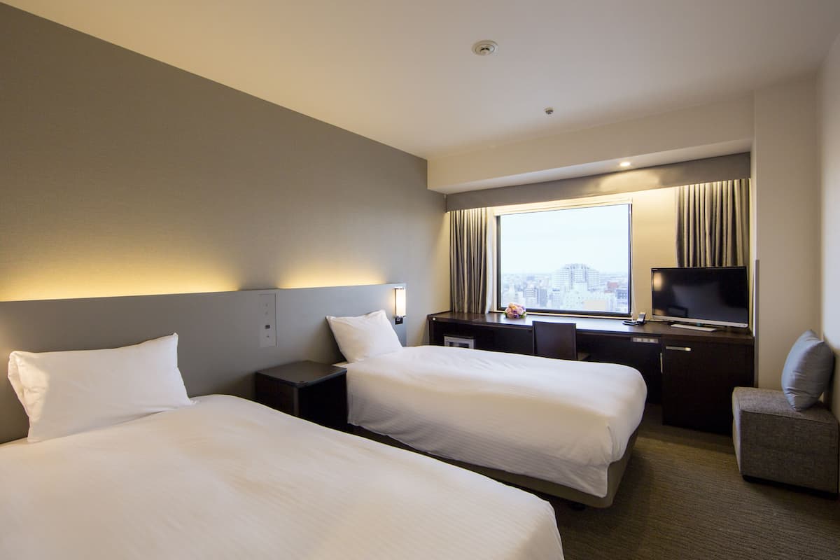 A hotel room with two beds, a desk with a television, a window with curtains, and a city view.