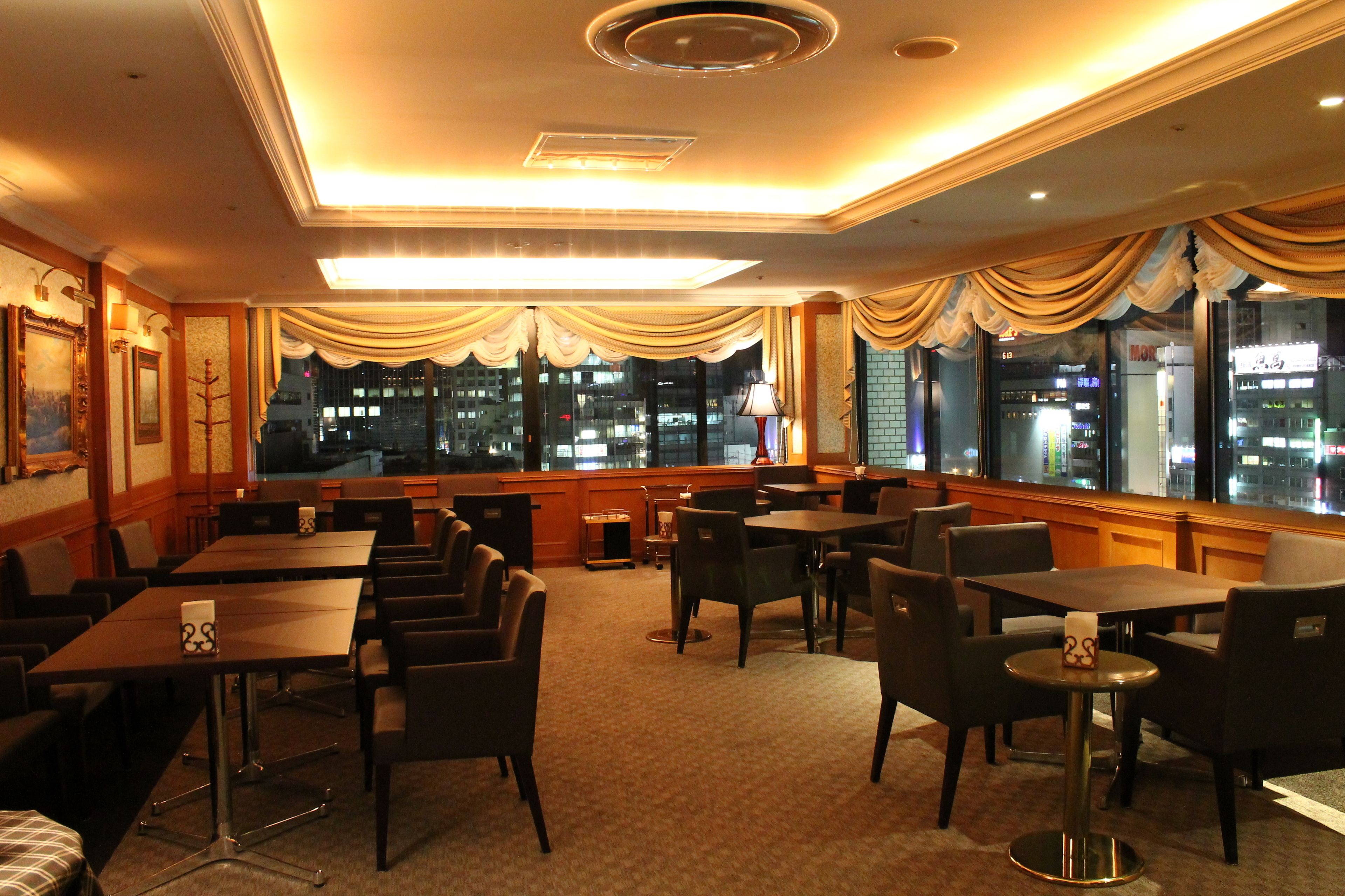 A hotel conference room with tables and chairs, large windows with curtains, and a view of the city at night.