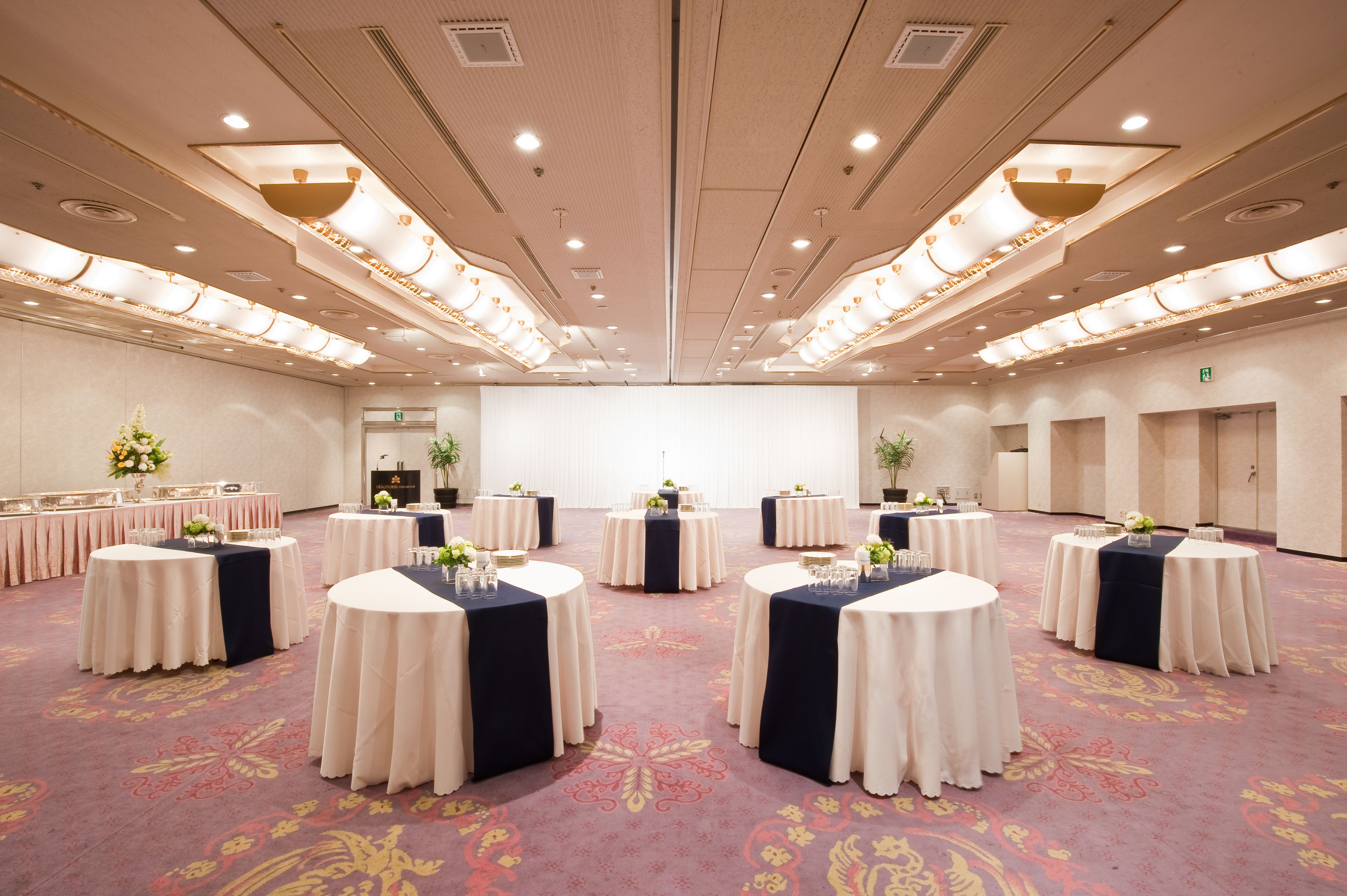 A large conference room with multiple round tables, each with a dark tablecloth and floral centerpieces. The room has a high ceiling with recessed lighting and a patterned carpet.