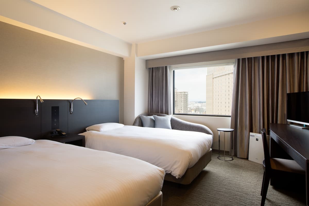 A hotel room with two beds, a desk, a chair, a TV, and a window with curtains.