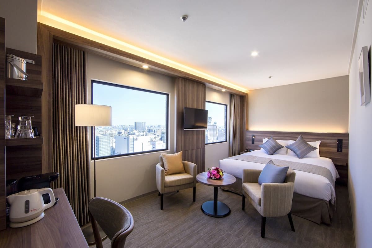 A hotel room with a large bed, two armchairs, a small table with a flower arrangement, a flat-screen TV, and a city view.