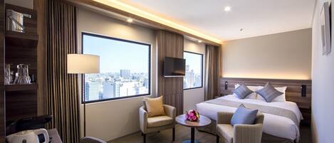 Habitación de hotel con una cama grande, dos sillones, una mesita con un arreglo floral, un televisor de pantalla plana y vistas a la ciudad.