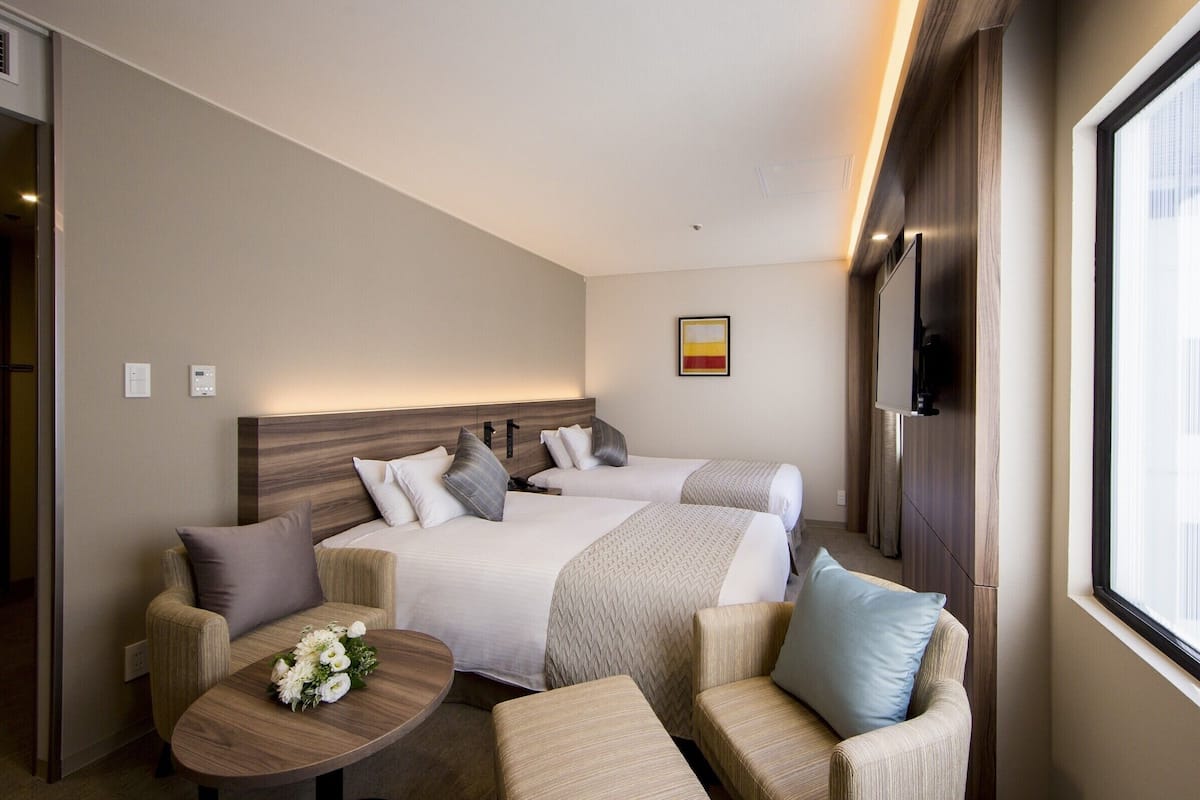 A hotel room with two beds, a sofa, a small table with flowers, and a TV mounted on the wall.