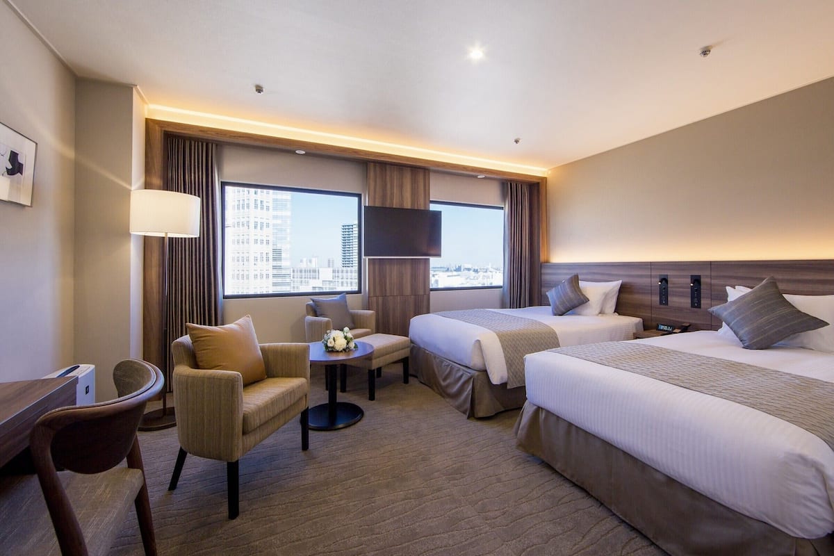 A hotel room with two beds, a desk, a chair, a small table with a flower arrangement, and a large window offering a city view.