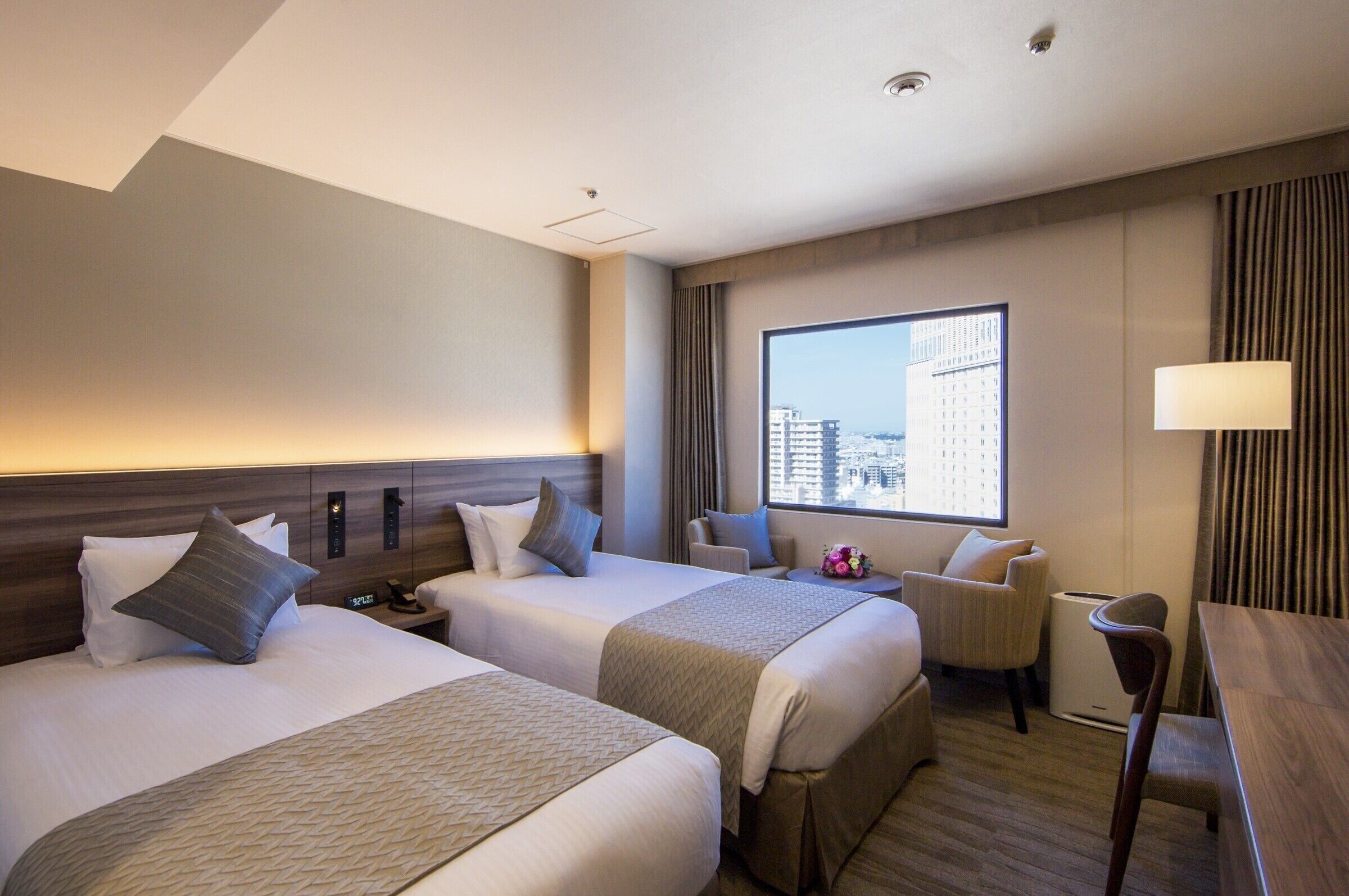 Une chambre d’hôtel avec deux lits, un bureau, une chaise, une fenêtre donnant sur la ville et une lampe.
