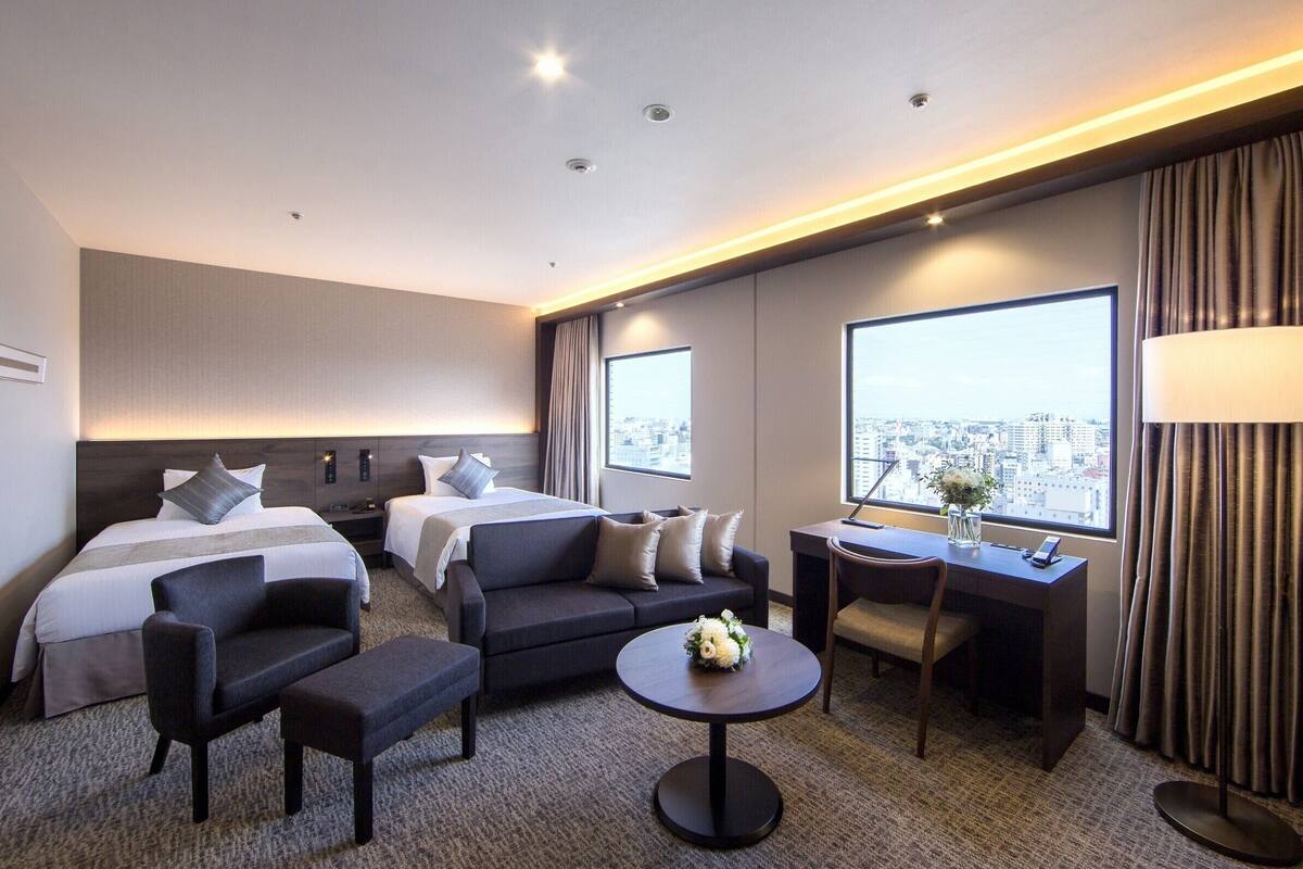 A hotel room with two beds, a sofa, a desk, and a view of the city.