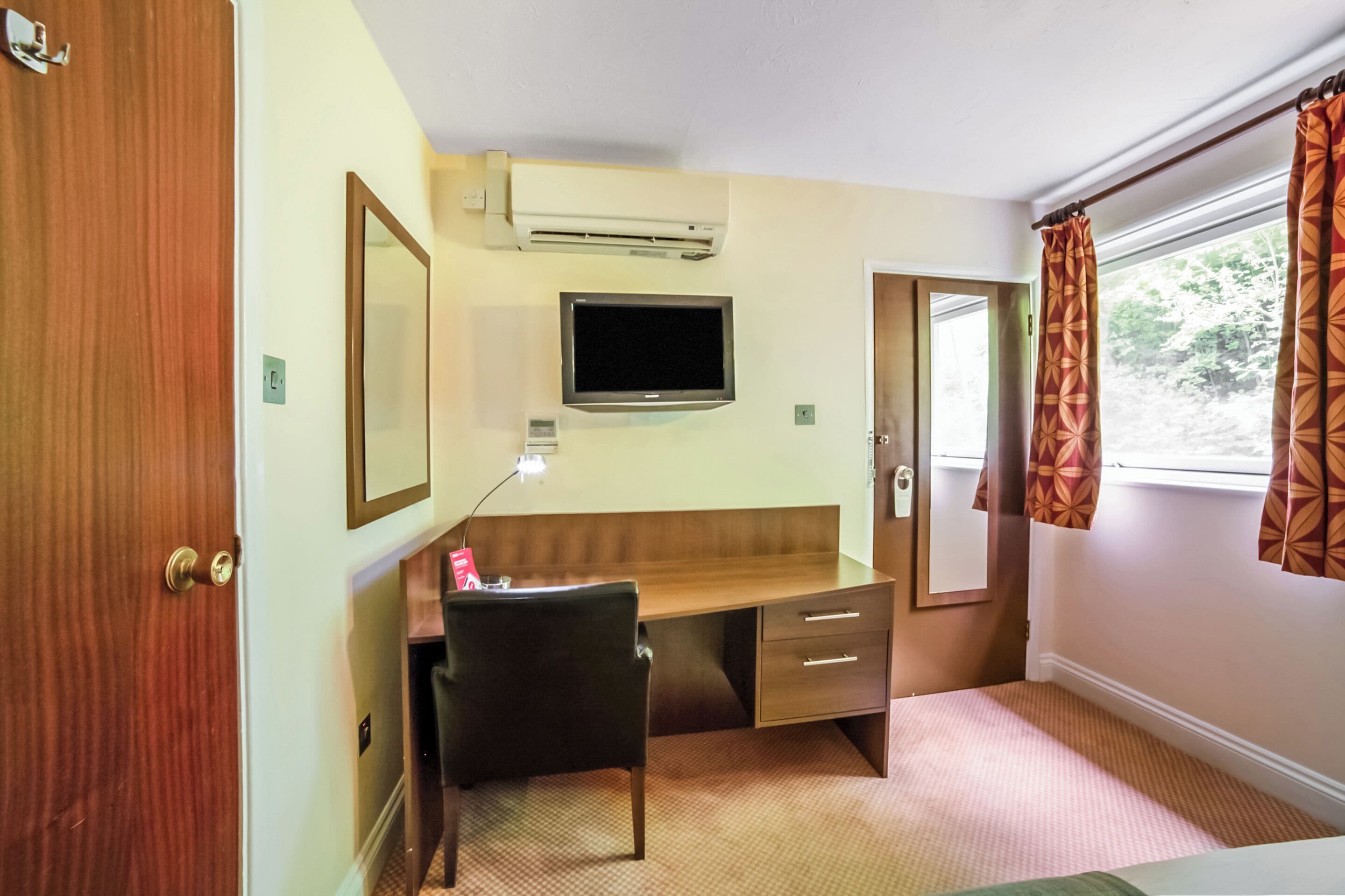 A hotel room with a wooden desk, a television, a mirror, and a window with patterned curtains.