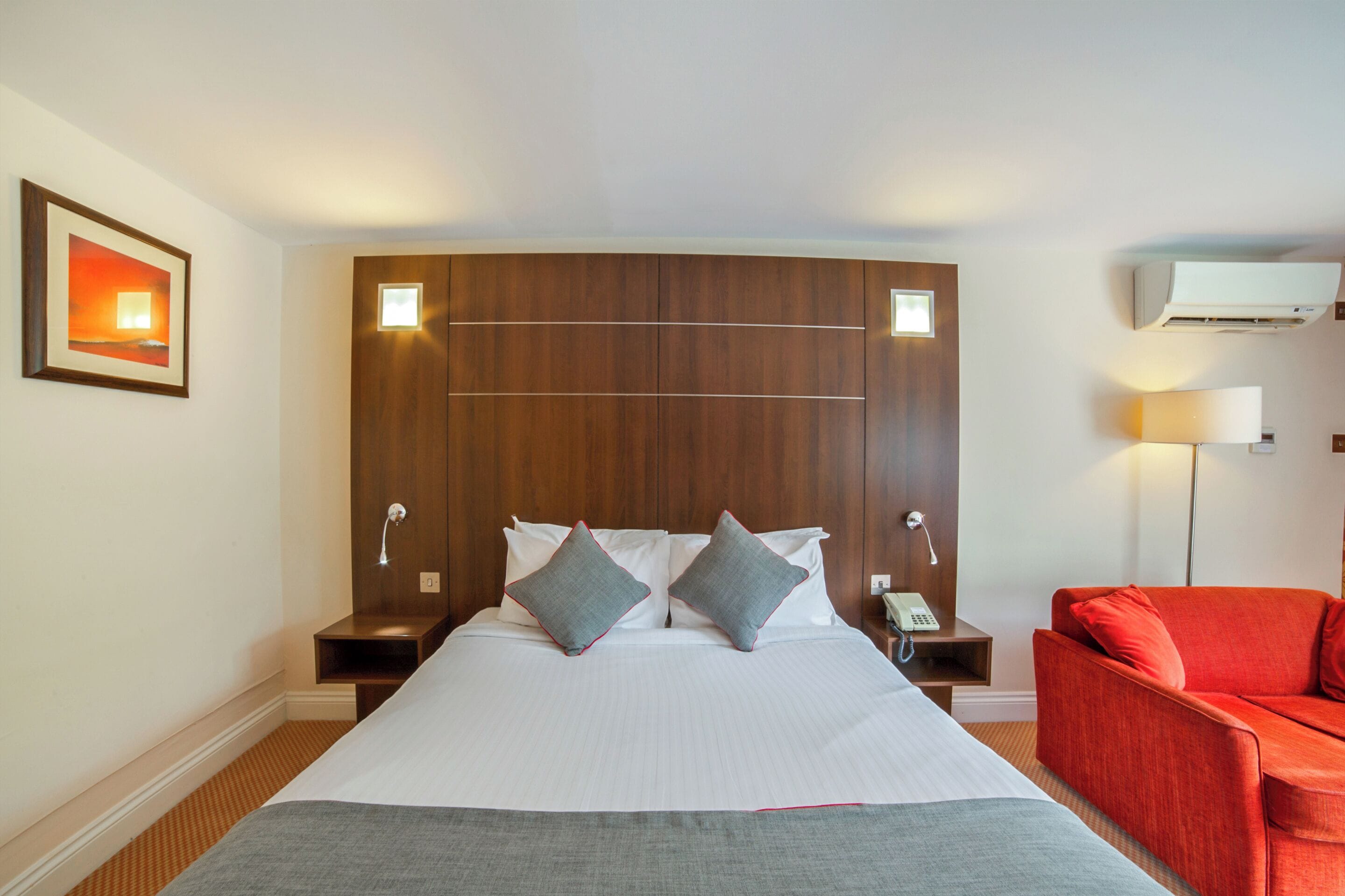 Habitación de hotel con una cama grande, dos mesitas de noche, un sillón rojo y un aire acondicionado de pared.