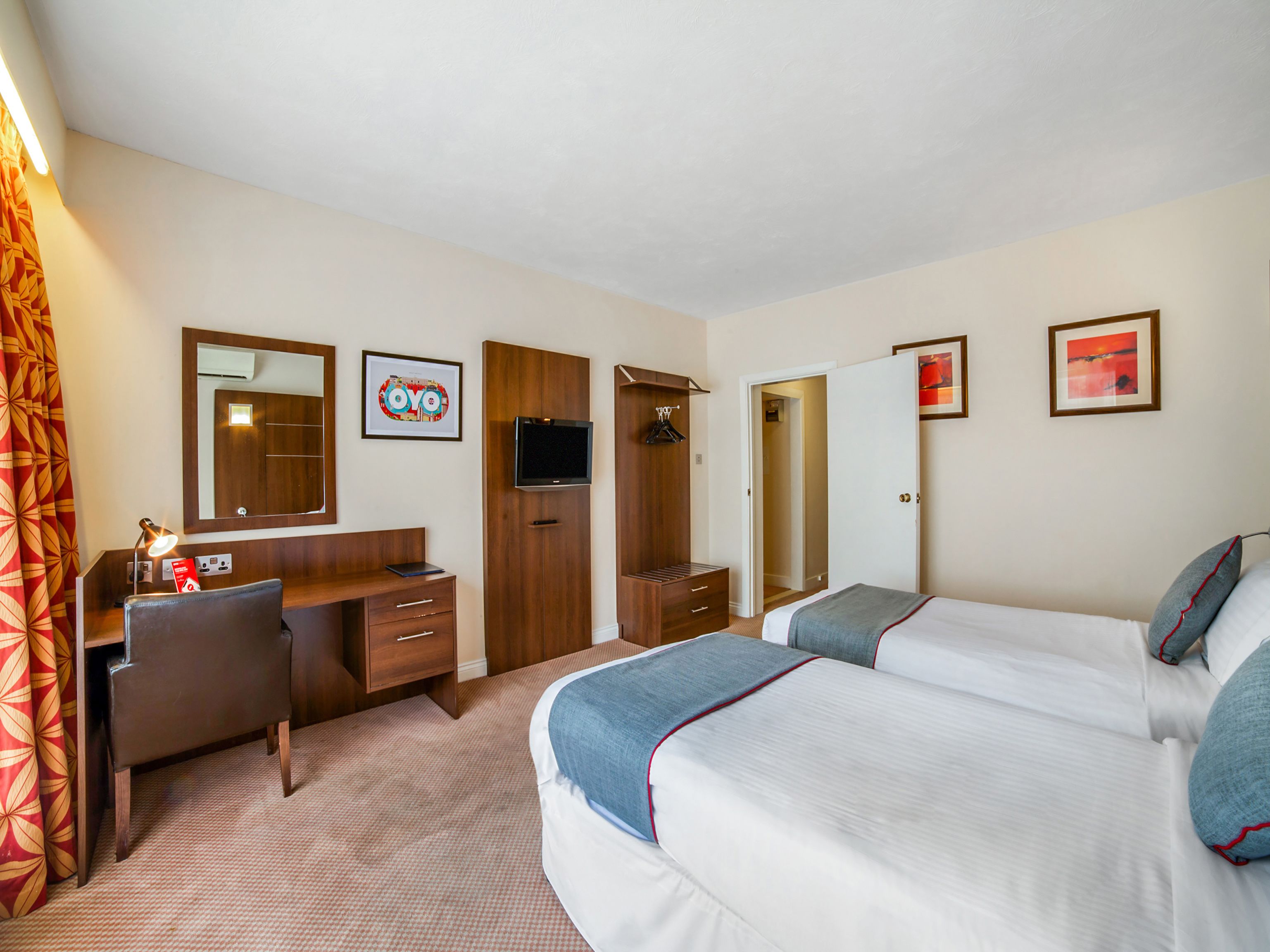 A hotel room with a desk, two beds, a wardrobe, a TV, and artwork on the walls.