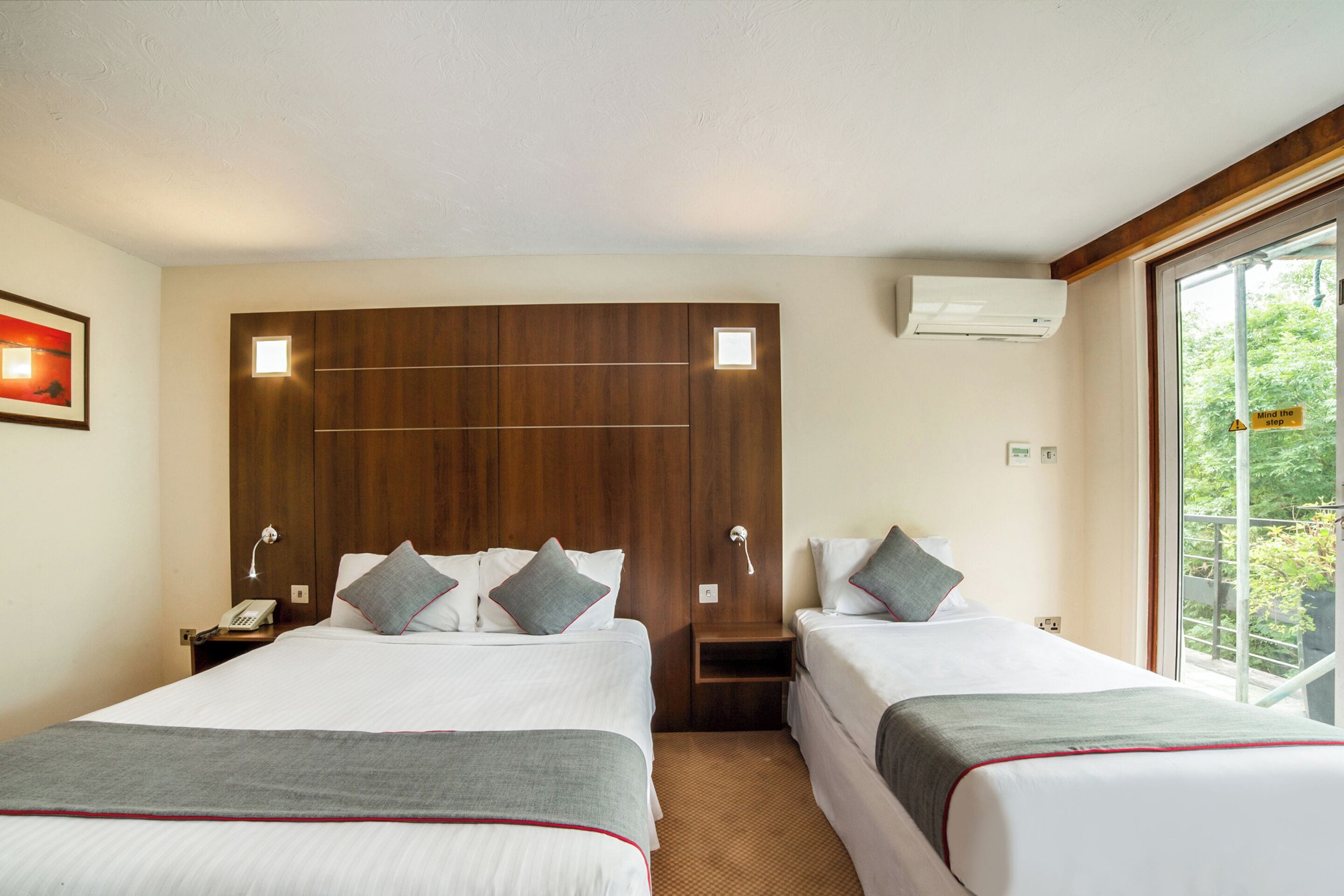 A hotel room with two beds, a wooden headboard, bedside lamps, a telephone, and a window with a view of trees.
