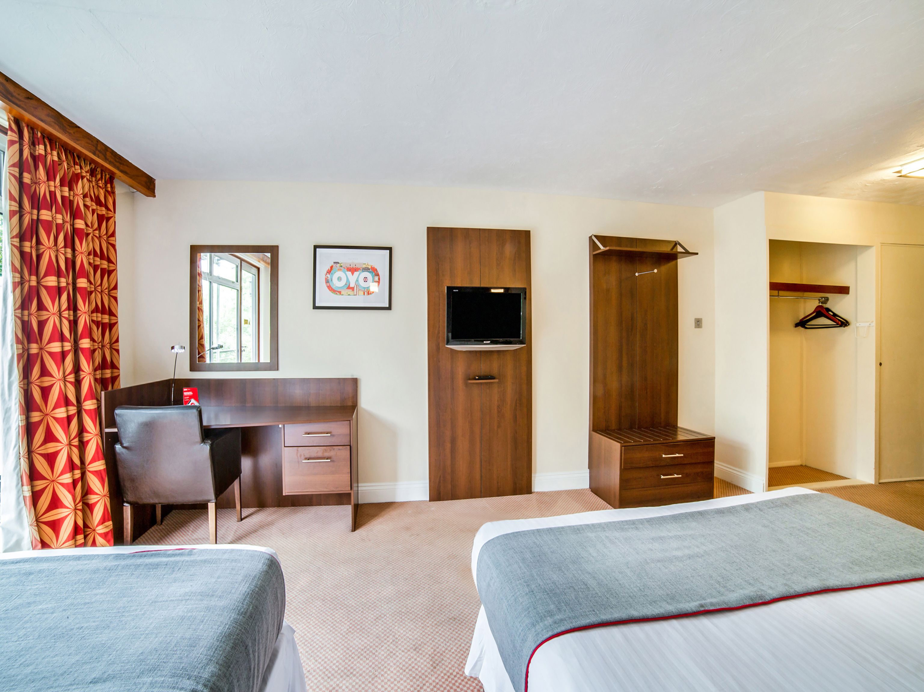 Quarto de hotel com duas camas, uma escrivaninha, um guarda-roupa, uma televisão e um espelho.