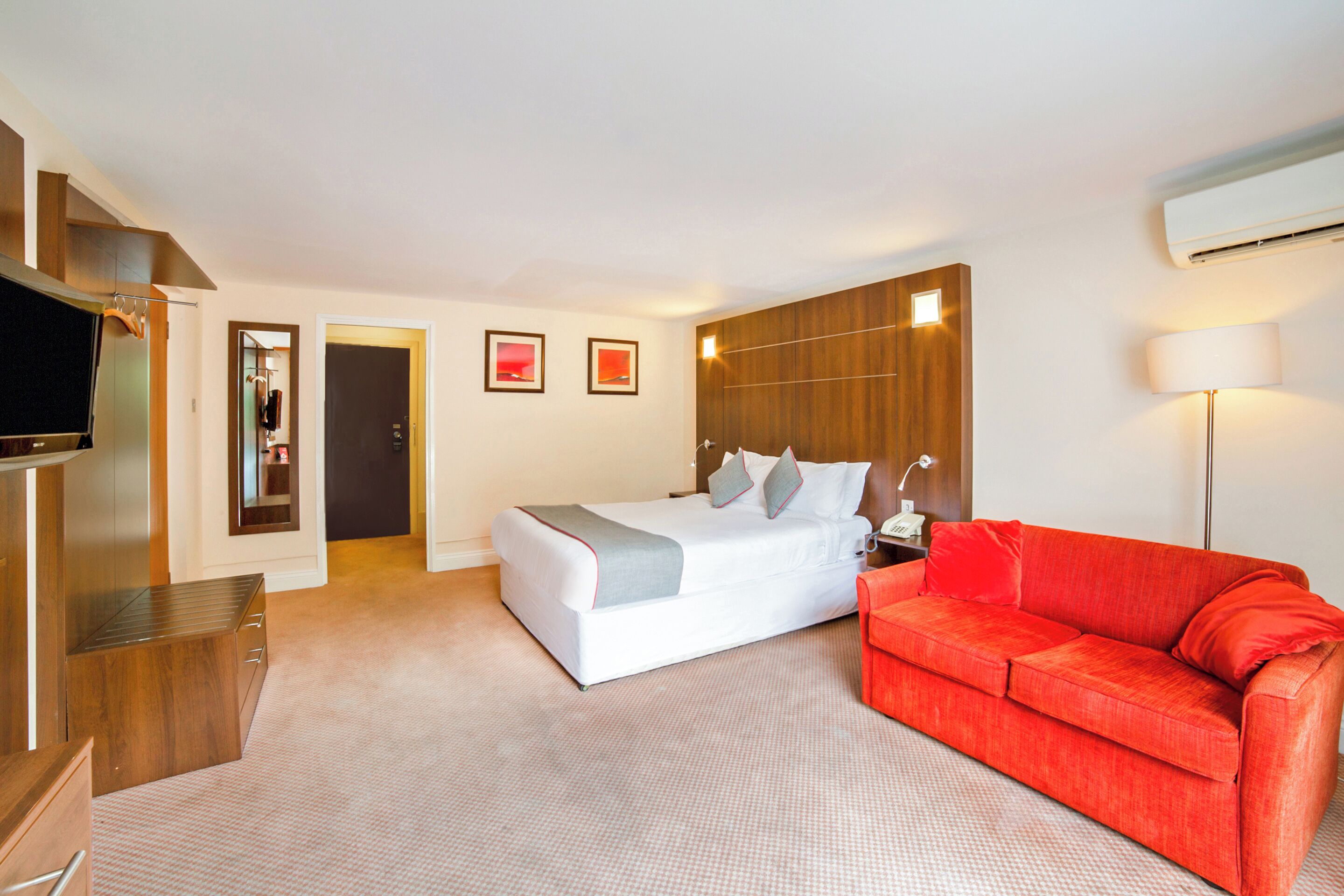 A hotel room with a bed, a red sofa, a TV, and a mirror.
