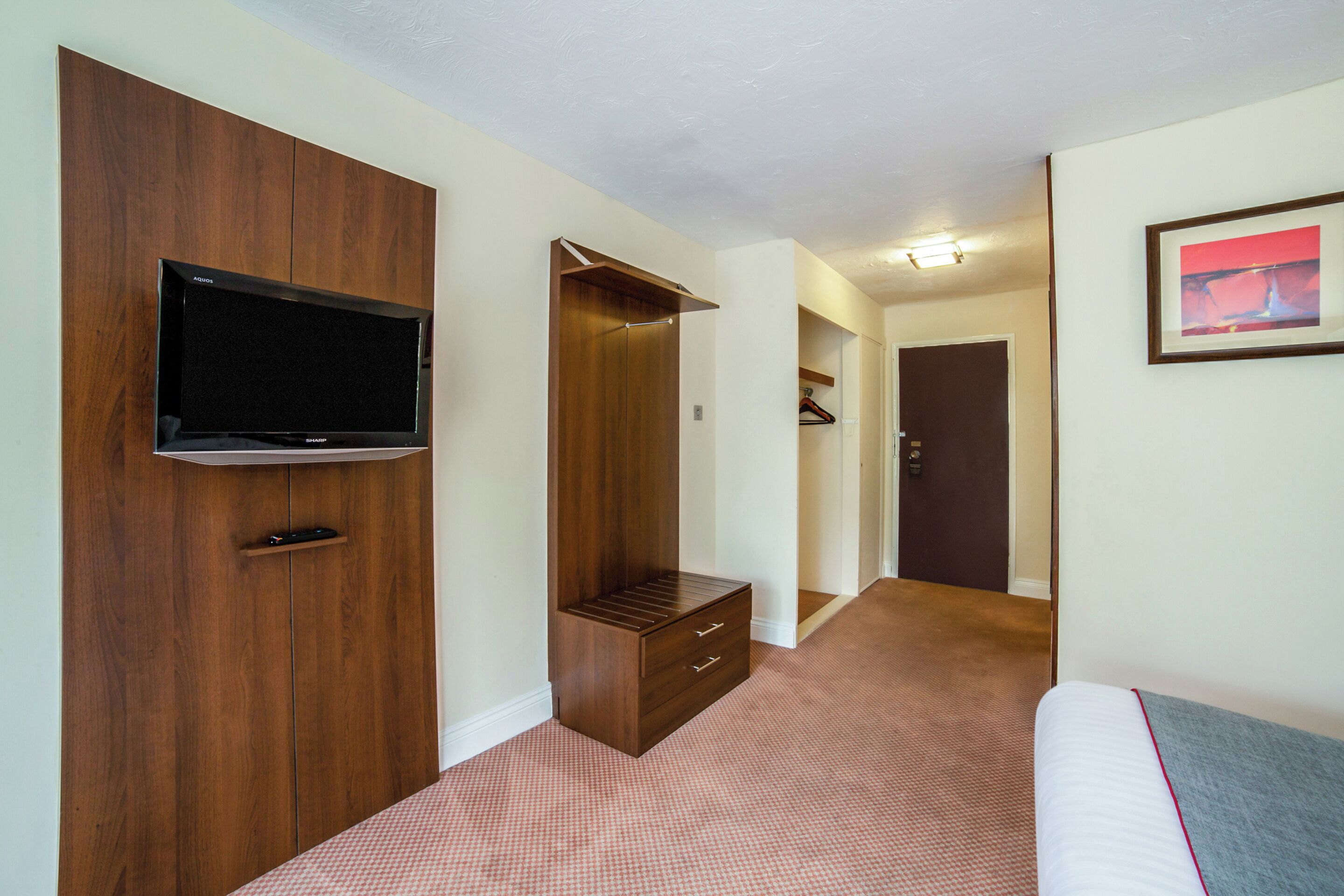 Habitación de hotel con un armario de madera, un televisor fijado en él, un escritorio pequeño y un cuadro enmarcado en la pared.