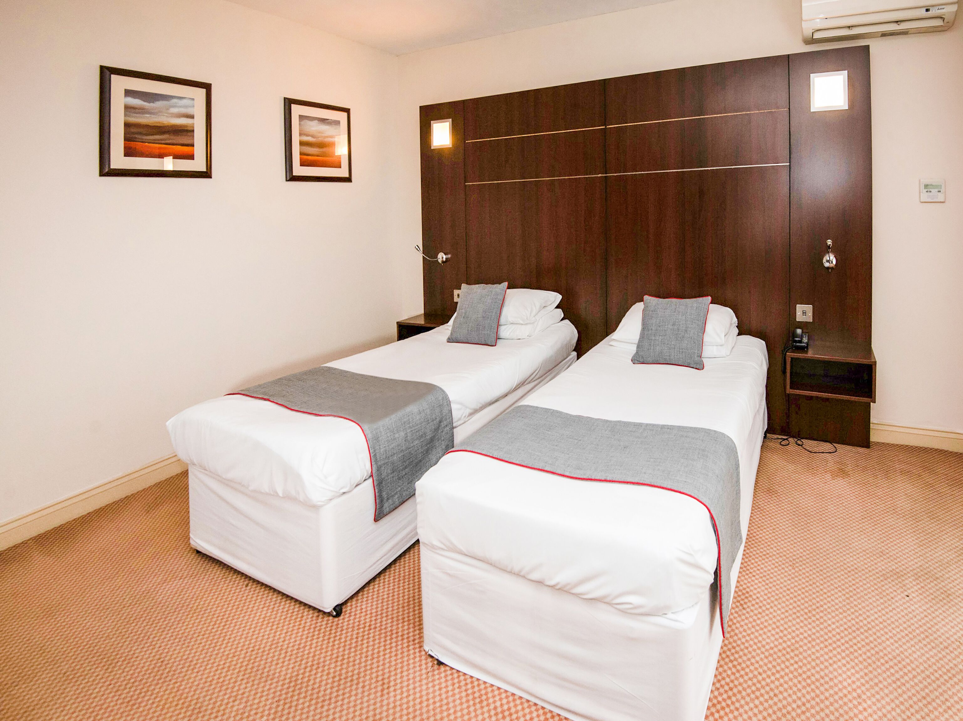 Habitación de hotel con dos camas individuales, cabecera de madera, dos cuadros enmarcados en la pared y una mesita de noche con una lámpara.