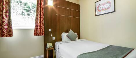A bedroom with a bed, a bedside table with a phone, a window with curtains, and a framed picture on the wall.