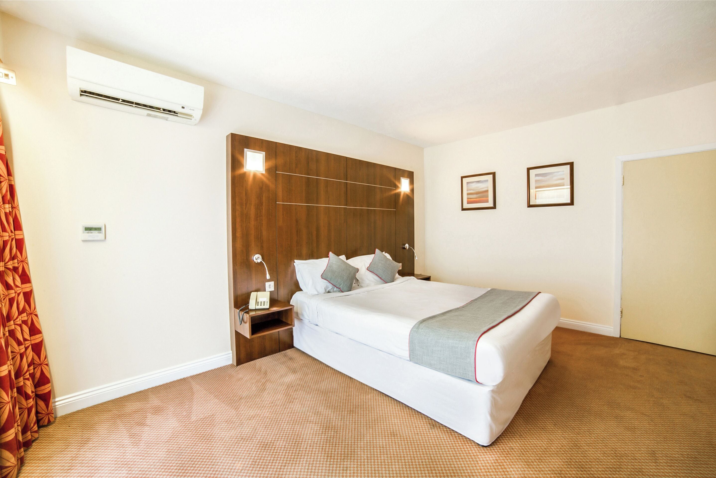A hotel room with a large bed, a wooden headboard, a nightstand with a lamp, and a wall-mounted air conditioner.