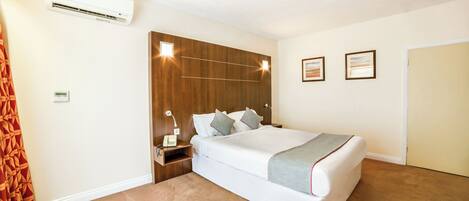 A hotel room with a large bed, a wooden headboard, a nightstand with a lamp, and a wall-mounted air conditioner.