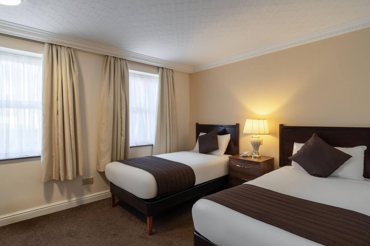 A hotel room with two beds, a nightstand with a lamp, and a window with curtains.
